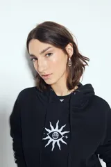 Canguro negro de calce relajado con capucha y cordones ajustables. Presenta un estampado gráfico frontal de un ojo con rayos de sol y la frase "STAY WEIRD".