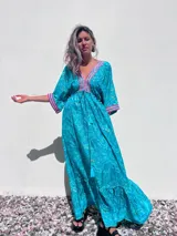 Túnica larga de estilo boho, color turquesa con estampado paisley en tonos azules y dorados. Presenta escote en V profundo con bordados decorativos en color fucsia y blanco, mangas amplias tipo kimono y ajuste en la cintura con cordón. El diseño incluye un ruedo con volado y terminaciones con borlas.