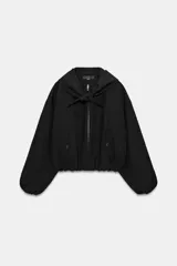 Chaqueta bomber negra con cuello redondo y pañuelo desmontable anudado al frente. Presenta cierre frontal con cremallera, bolsillos laterales con solapa y botones a presión, y terminaciones elásticas abullonadas en puños y ruedo.