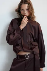 Cazadora satinada de color negro, con cuello subido, manga larga y bolsillos frontales de plastrón con cordones decorativos. Cuenta con bajo elástico y cierre frontal con cremallera y botones a presión oculta.