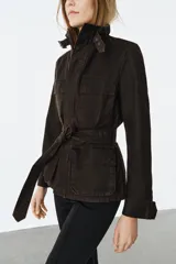 Chaqueta de estilo safari confeccionada en mezcla de algodón con acabado encerado. Presenta cuello alto con trabilla ajustable y hebilla metálica, cierre frontal con cremallera y botones ocultos bajo solapa, bolsillos de plastrón con solapa en el delantero y cinturón ajustable del mismo tejido para anudar en la cintura.