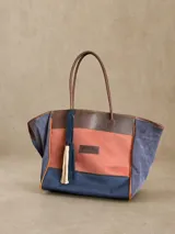 Bolso tipo shopper de gran tamaño, confeccionado en una combinación de lona y gamuza. Presenta un diseño tricolor con paneles en negro, blanco y marrón, con detalles en cuero marrón y borlas decorativas colgantes. Incluye doble asa de hombro y cierre principal.