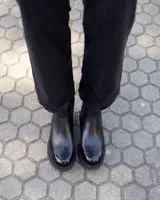 Botinetas tipo Chelsea de cuero negro, con paneles laterales elásticos y suela track dentada de goma. Presentan una combinación de texturas entre cuero mate y cuero charolado en la puntera y el talón.
