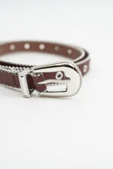 Cinturón color bordeaux con detalle de tachas metálicas en los bordes y ojales metálicos a lo largo de toda la tira. Incluye hebilla metálica plateada con diseño labrado.