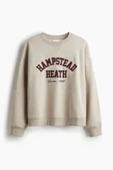 Buzo oversized color beige con interior cepillado suave. Presenta hombros caídos y un diseño de texto en el pecho que dice "HAMPSTEAD HEATH London, 1998" en rojo y negro. Tiene ribete acanalado en cuello, puños y bajo.