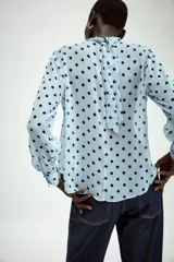Camisa de manga larga con cuello fluido y diseño cruzado en el frente. Confeccionada en tejido semitransparente de color celeste con estampado de lunares negros. Presenta bajo asimétrico y cierre en la espalda con botones forrados.