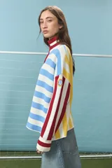 Sweater de punto de algodón con diseño de bloques de rayas verticales y horizontales en colores rojo, amarillo, azul y blanco. Presenta cuello alto con cierre frontal parcial y mangas largas con puños acanalados.