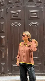 Camisa de color marrón chocolate con cuello redondo grande y ribetes en color amarillo vibrante. Presenta cierre frontal con botones amarillos, mangas largas con puños elásticos y un corte holgado.