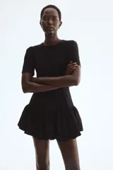 Vestido corto negro con escote redondo, mangas cortas abullonadas y cierre frontal de botones forrados. Cuenta con costuras moldeadoras en la cintura y falda con bajo globo fruncido.