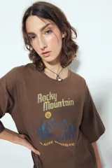 Remera marrón de manga corta con cuello redondo y calce relajado. Presenta un estampado frontal con la frase "Rocky Mountain National Park" en letras doradas y azules, junto a un paisaje montañoso.