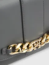 Bandolera gris con solapa y cierre a presión. Presenta una correa ajustable y una cadena dorada gruesa en la parte delantera con el monograma TH.