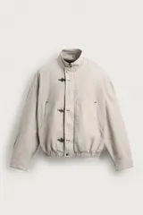 Cazadora relaxed fit confeccionada en tejido de algodón de color beige, con cuello subido y manga larga. Cuenta con bolsillos de plastrón en la cadera, bajo elástico y cierre frontal con cremallera oculta y alamares metálicos.