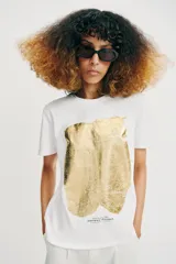 Camiseta de algodón con cuello redondo y manga corta. Presenta un diseño estampado frontal con efecto metalizado plateado sobre fondo rectangular.
