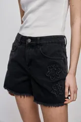 Short de denim blanco de calce recto y tiro medio, con bolsillos delanteros, cierre con botón y terminación desflecada en el ruedo. Presenta detalles florales bordados al tono en el frente.