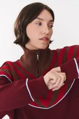 Buzo de tejido de punto en color azul marino, con cuello alto y cierre frontal parcial. Presenta un diseño con franjas en color marrón y detalles de líneas en rojo en el pecho, hombros y ruedo. Tiene corte holgado y mangas largas con puños acanalados.