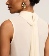 Blusa color crema sin mangas de crepé con cuello alto y lazo por tiras largas de tejido. La prenda tiene cierre por gota con detalle de perla artificial atrás del cuello y frunzidos en la parte delantera.