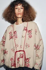 Chaqueta acolchada color beige con bordados florales en tonos rosados y rojos. Tiene escote en pico, manga larga y forro interior rojo. El cierre frontal es con alamar y tiene ribetes rojos.