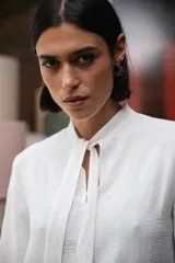 Blusa blanca de textura arrugada, con cuello alto, abertura frontal tipo gota y mangas largas acampanadas con detalle de frunces.