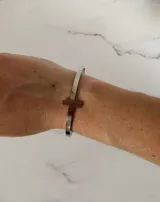 Brazalete tipo esclava rígida abierta, fabricada en acero quirúrgico, con un detalle de cruz integrada en la estructura.
