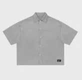 Sobre camisa de gabardina en color gris, con corte boxy fit, cuello clásico, cierre frontal con botones y etiqueta decorativa en el ruedo.