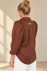 Camisa de lino color blanca, con cuello clásico, mangas tres cuartos abullonadas en los hombros y terminación con volados en los puños, cierre frontal con botones ocultos.