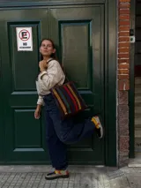 Cartera tipo tote de cuero con diseño de franjas verticales multicolores en tonos tierra, rojo y azul. Cuenta con correas trenzadas de cuero y cierre superior.