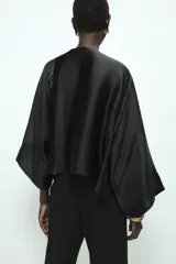 Blusa negra de satén con escote fluido, diseño drapeado en el frente y mangas largas anchas. Presenta un bajo asimétrico y una caída suave y elegante.