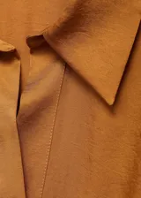 Camisa de tejido satinado en color mostaza, con diseño cruzado, cuello camisero, manga larga con puños abotonados y cierre delantero de botones ocultos.