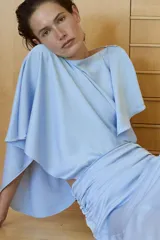 Vestido midi de tejido satinado color celeste, con cuello barco y diseño asimétrico con mangas tipo capa. Presenta un detalle drapeado en la cintura y abertura en la espalda.