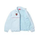Campera Supreme de nylon color celeste, con cuello alto, cierre frontal y una línea horizontal roja en el pecho. Cuenta con escudo bordado y puños y ruedo elásticos.