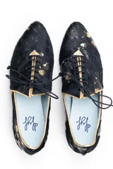 Zapato tipo blucher de pelo de pony en color negro con salpicaduras doradas. Presenta cierre de cordones, ribetes dorados a contraste y una silueta geométrica con aberturas laterales. Cuenta con un tacón bajo forrado en el mismo material.