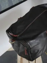 Bolso de viaje tipo weekender confeccionado en cuero negro, con manijas y correa ajustable en cuero color bordó. Presenta cierre metálico frontal con detalle en azul, bolsillo externo con cierre y parche de marca grabado en el frente.