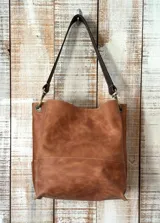 Cartera tipo bolso de cuero color marrón, con diseño estructurado, costuras visibles y correa de hombro en tono oscuro. Cuenta con herrajes metálicos en color bronce.