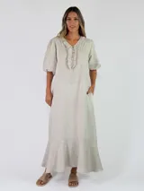 Vestido largo de lino color beige, con escote en V rodeado de volados, mangas cortas abullonadas, bolsillos laterales y falda con volado en el ruedo.