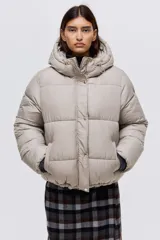 Campera puffer color beige con capucha, cierre frontal y solapa con botones a presión. Cuenta con bolsillos laterales, elástico en los puños y cordón ajustable en la capucha y el ruedo.