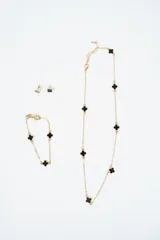Set de joyería compuesto por un collar largo, una pulsera y un par de aros, todos con diseño de tréboles negros con borde dorado. El collar y la pulsera presentan una cadena fina de eslabones dorados intercalados con los tréboles.