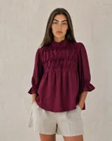 Blusa holgada color bordó con detalle de nido de abeja en el canesú delantero, cuello alto con volados y mangas tres cuartos con puños elásticos fruncidos.