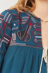 Vestido amplio color azul petróleo con bordados étnicos en tonos rojo, blanco y azul oscuro en el canesú y mangas. Presenta escote en V con cordones de ajuste terminados en borlas rojas y doradas.