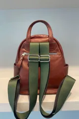 Mochila de cuero vacuno color tostado, con detalles en cuero metalizado plateado en los laterales y manija superior. Presenta un bolsillo frontal con cierre metálico, correas de cinta regulables y placa metálica con logo de la marca en la parte inferior.