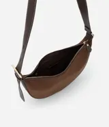 Cartera bandolera de formato media luna, confeccionada en material sintético efecto gamuza color marrón. Presenta correa larga ajustable con costuras a la vista y cierre superior con cremallera.