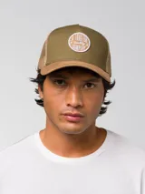 Gorro trucker de color verde militar, con visera curvada marrón claro, panel frontal rígido y red trasera. Cuenta con un parche circular bordado en la parte delantera y cierre ajustable snapback.
