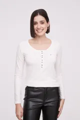 Remera blanca de manga larga, entallada, con cuello redondo y una hilera de botones en el frente. Incluye un pequeño bolsillo decorativo en el pecho.