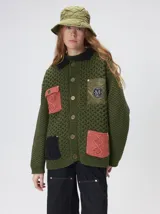 Campera tejido grueso y abrigado color verde militar, con forro de nylon interno, cuello estilo camisa en color negro y cierre frontal con botones. Cuenta con cuatro bolsillos delanteros con apliques en distintos tonos y texturas, y un parche bordado circular en el bolsillo superior izquierdo.