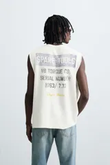 Camiseta sin mangas (manga sisa) de punto color crema, corte relaxed fit, con cuello redondo y acabados irregulares en las sisas. Presenta un texto estampado en el pecho que dice "SERIAL N. 8763/ 7.3Z".