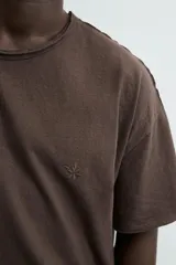 Camiseta de corte relajado (relaxed fit) color blanco roto, con cuello redondo y manga corta. Presenta un bordado sutil en el pecho con diseño floral en tonos claros.