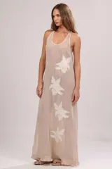 Vestido maxi de lino color beige, con textura calada, escote redondo y espalda deportiva. Presenta detalles de grandes flores bordadas blancas a lo largo de la prenda y terminación de flecos en el bajo.