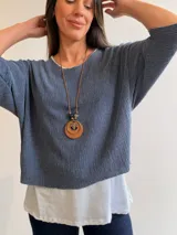 Conjunto de dos piezas compuesto por una remera básica blanca de algodón y un buzo liviano de textura plisada en color azul grisáceo. El buzo presenta un diseño de manga murciélago, corte holgado y detalles de hilo plateado en los bordes. Incluye un collar largo con dije circular de madera y metal.