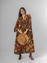 Vestido largo de manga larga, corte oversized y cuello camisero con abertura en V y botones. Presenta un estampado botánico en tonos tierra sobre fondo marrón oscuro, con volados en el ruedo.