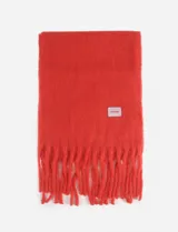 Bufanda de textura suave y acabado afelpado, color negro, con flecos en los extremos y un pequeño parche cuadrado decorativo con logo en color verde y azul.