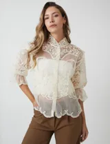Blusa de tul bordado color beige, marca Liv Los Angeles, con cuello alto, cierre frontal de botones y mangas ¾ amplias con volados.